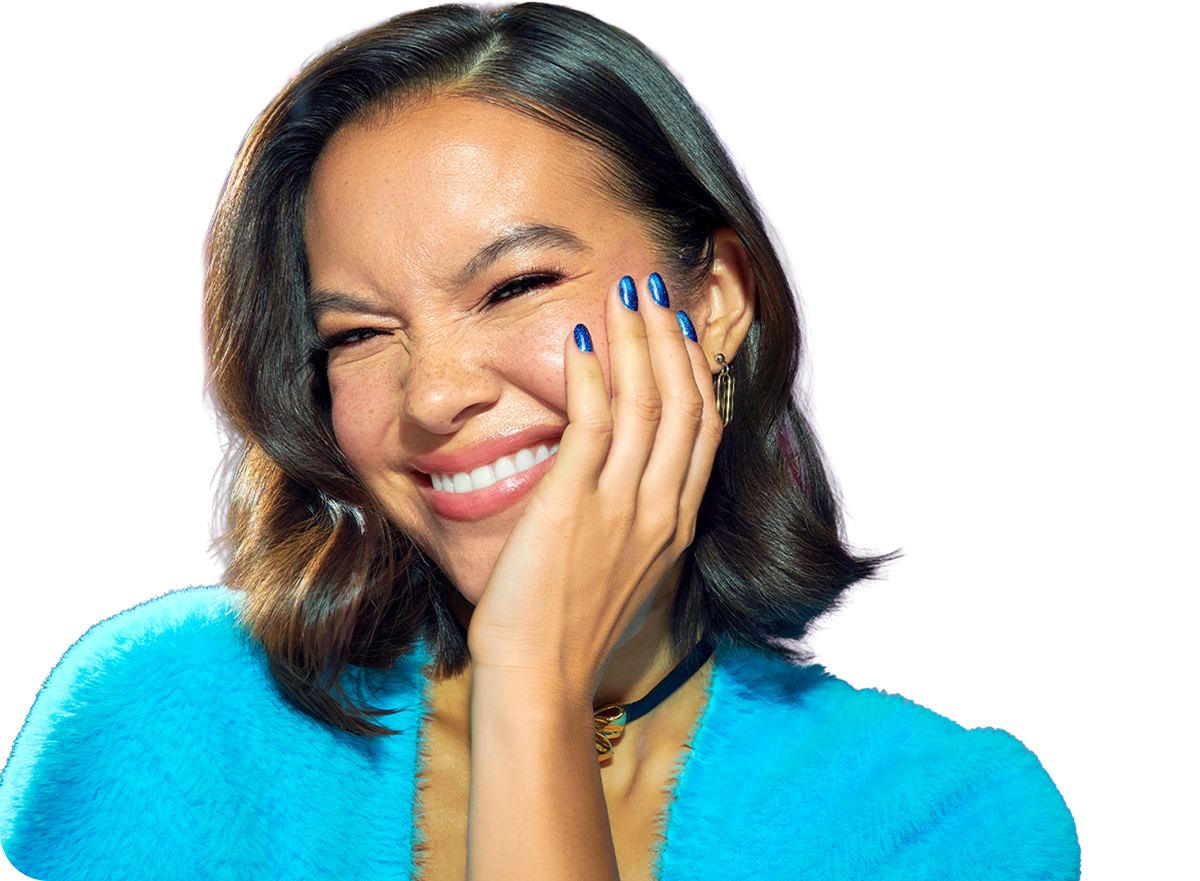 A close-up portrait of a woman with dark shoulder-length hair, smiling broadly and covering part of her face with her hand. She is wearing a bright turquoise fuzzy top against a plain white background.