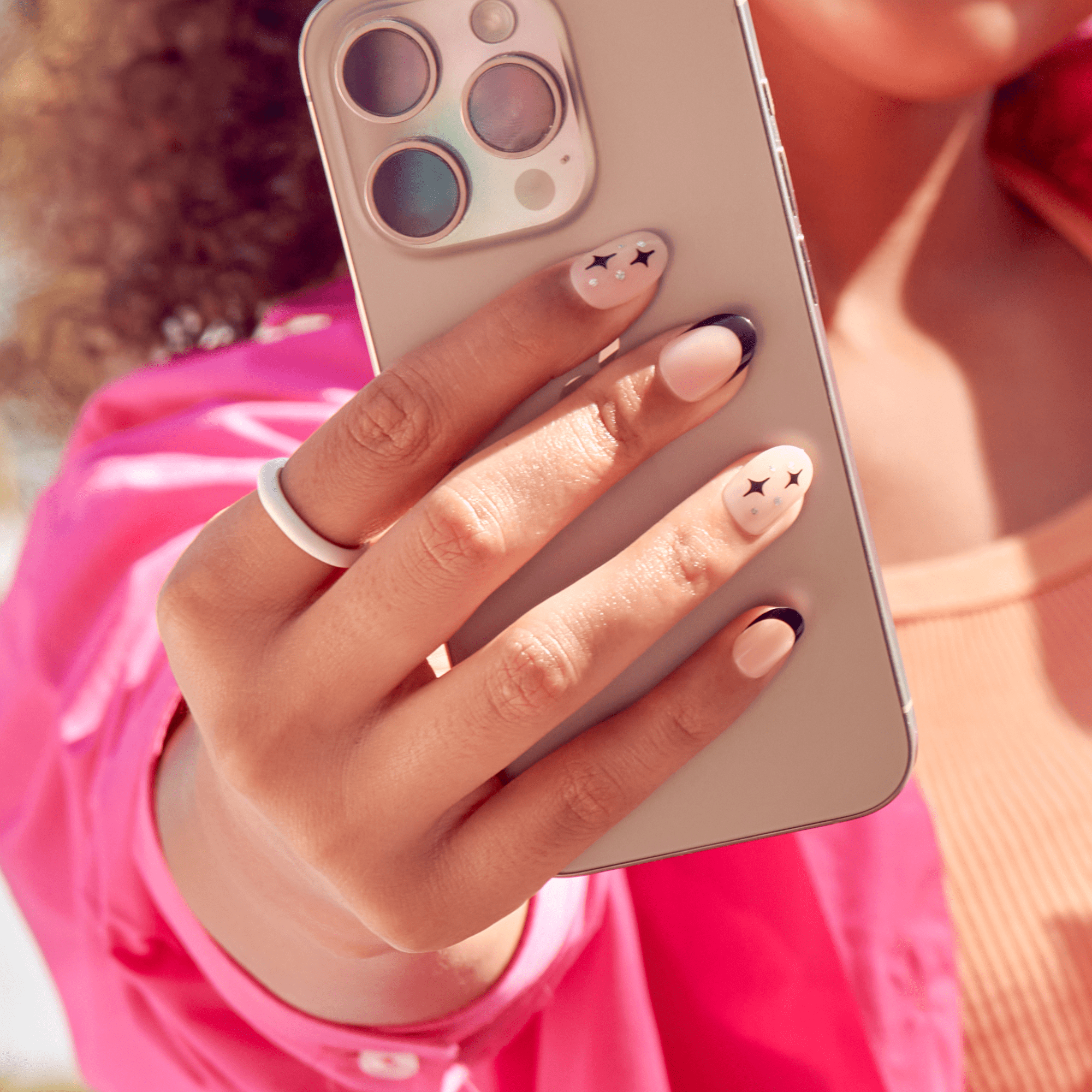 Hand holding a silver smartphone showing press-on nails in soft pink with black star designs and black French tips, worn with a white ring and pink clothing.