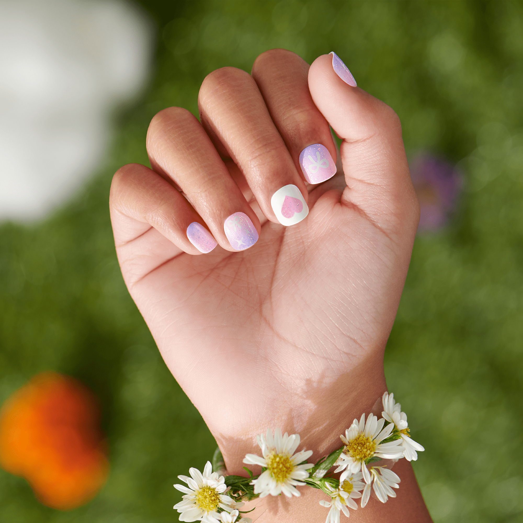 Hand with lavender and white nail art, wearing daisy flower bracelet against green garden background.