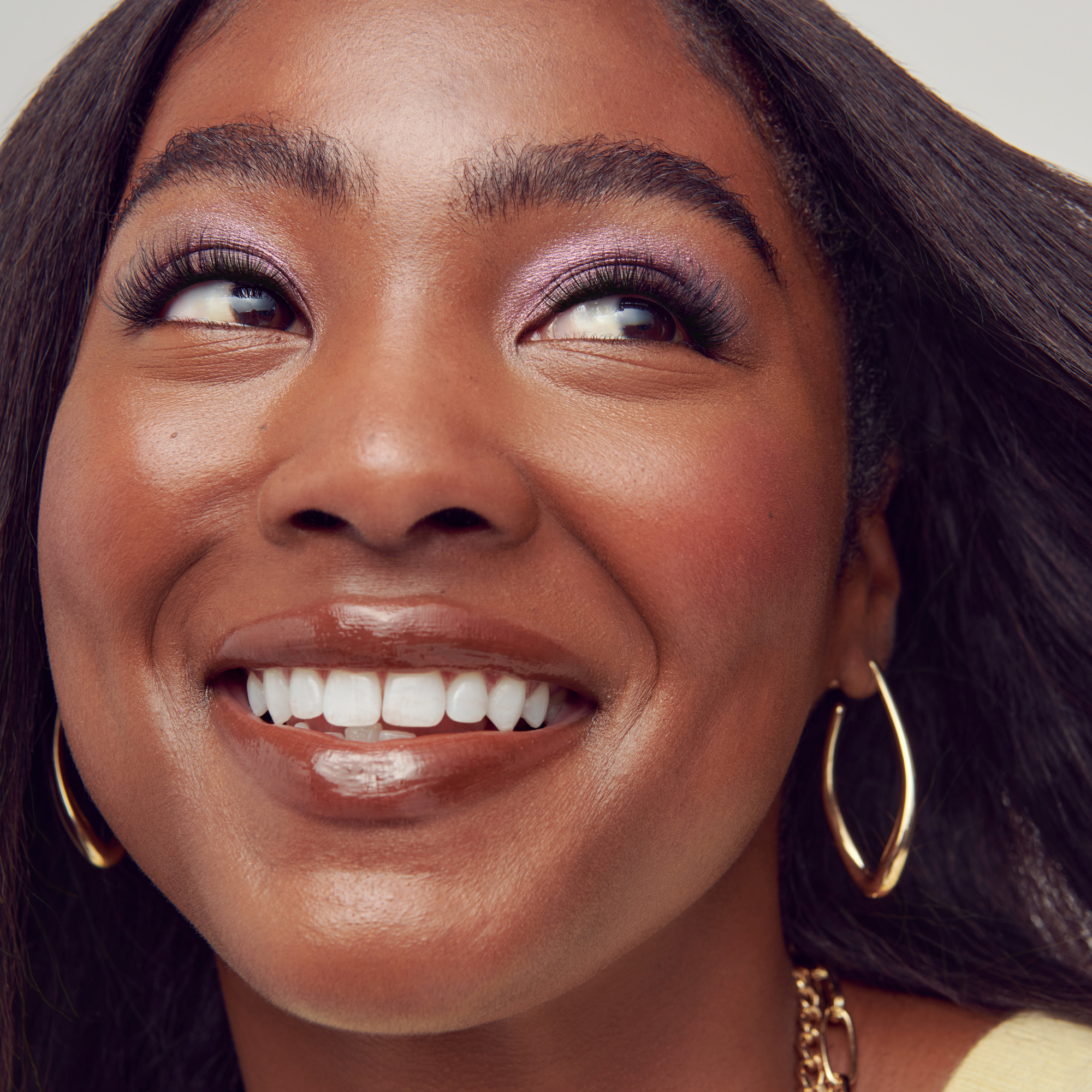 Woman with dark hair smiling, wearing gold hoop earrings and purple eyeshadow.