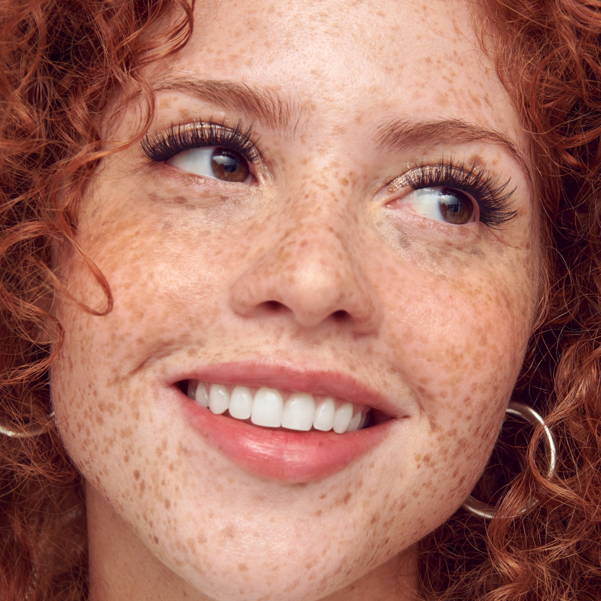 Close-up of a person with red curly hair and freckles, wearing a hoop earring.