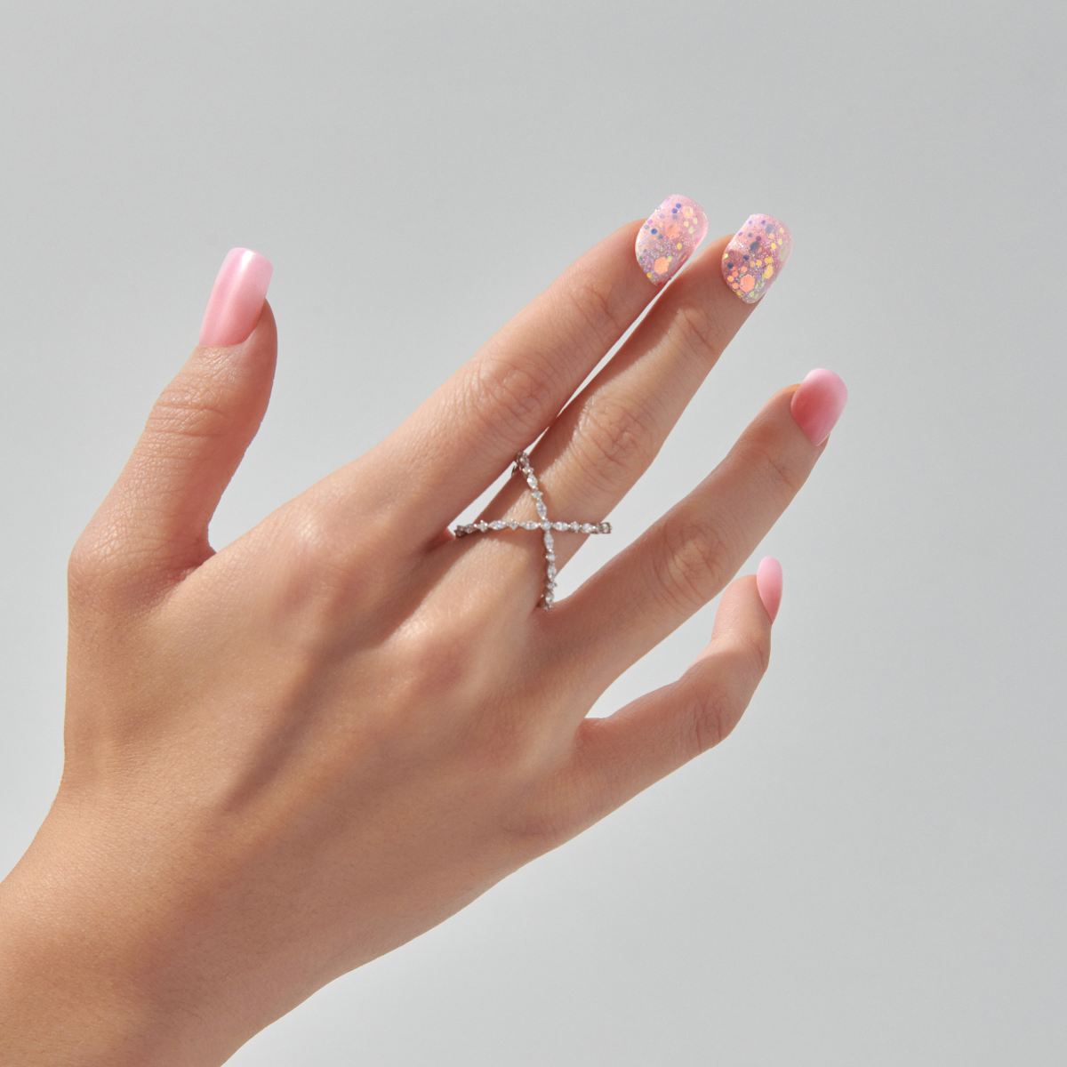 Hand with pink nail polish and glittery accent nails, wearing diamond cross ring.