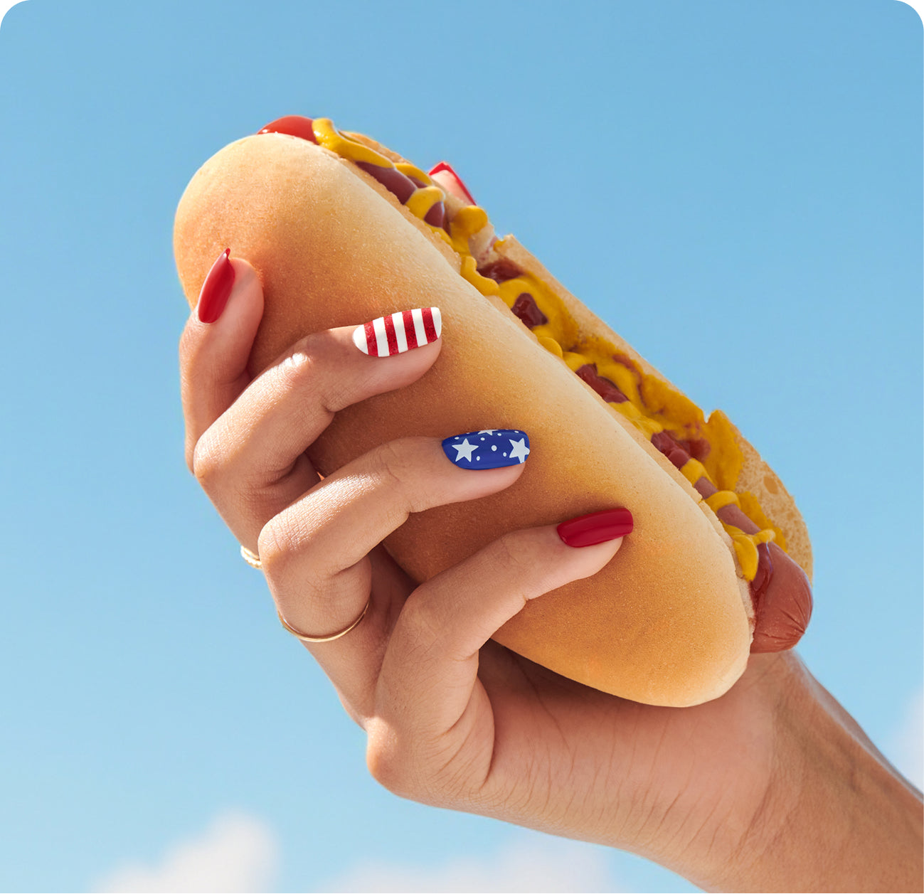 Hand holding hot dog with patriotic nail art against blue sky.