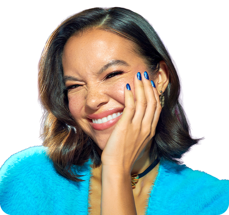 A close-up portrait of a woman with dark brown hair, a bright blue fuzzy top, and blue nail polish, smiling and touching her face against a plain white background.