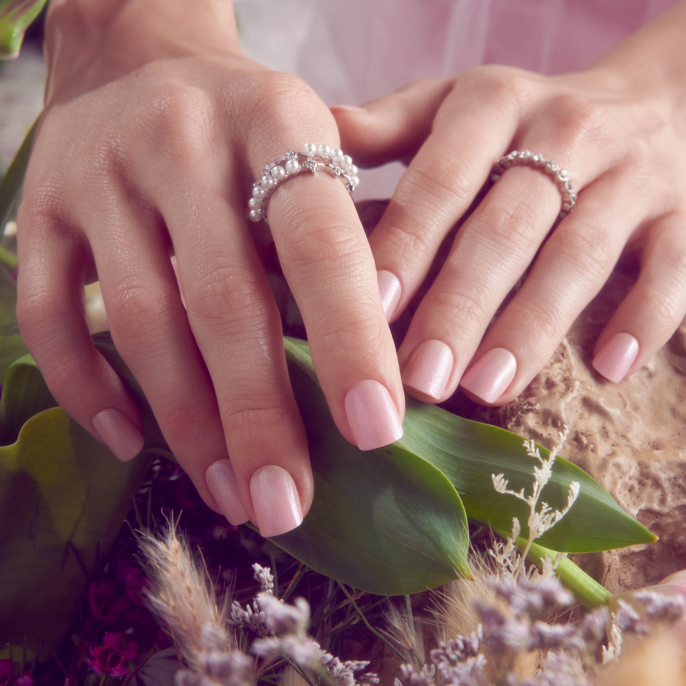 Two hands with pearl rings and pink nail polish on flowers and greenery.