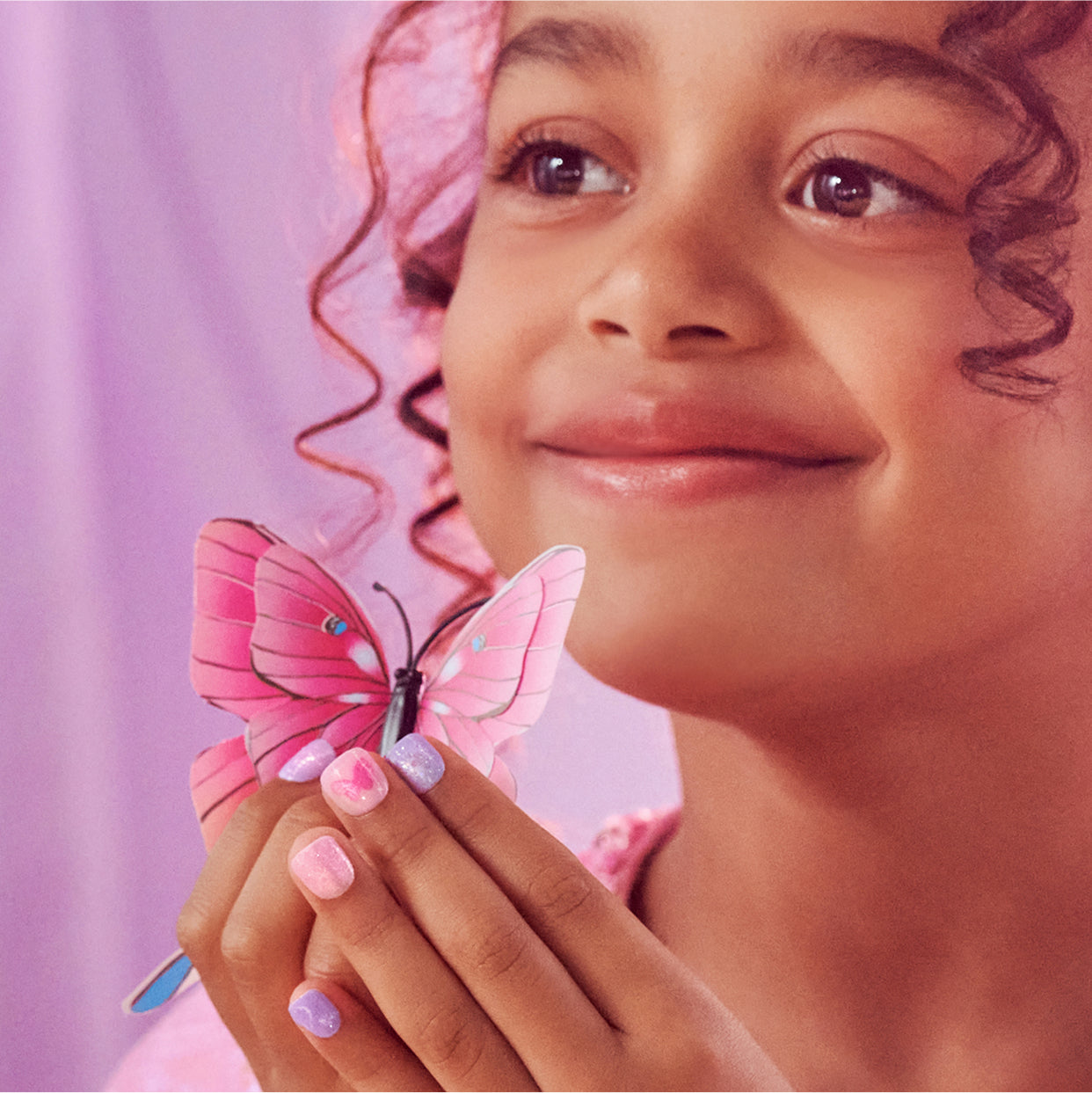 Child holding pink butterfly with manicured nails against pink background.
