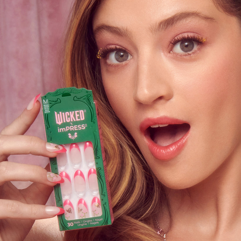 Woman holding Wicked imPRESS press-on nails with pink and white design against pink background.