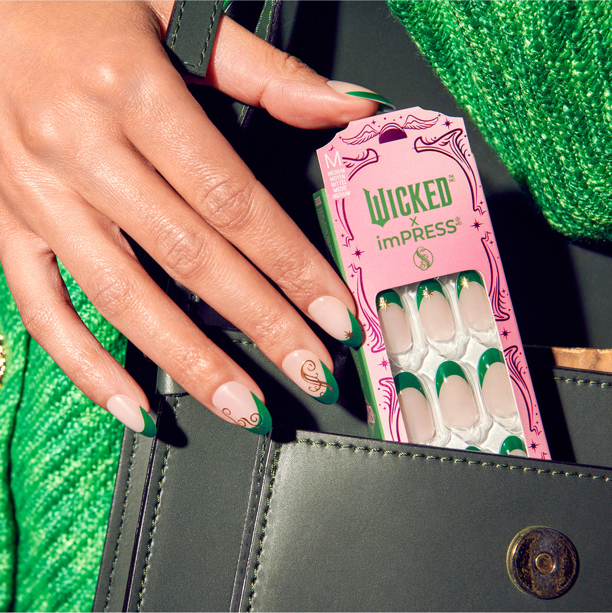 Hand displaying Wicked x imPRESS press-on nails with green and white design next to pink packaging.