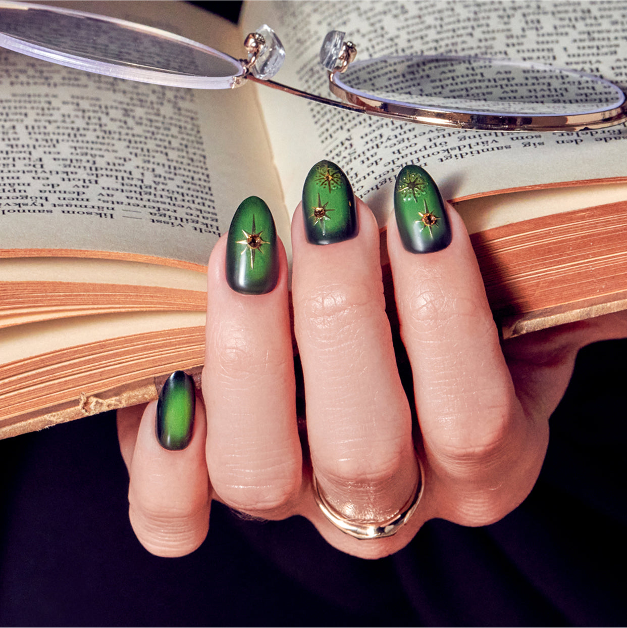 Green and gold celestial nail art with star designs on open books and eyeglasses backdrop.