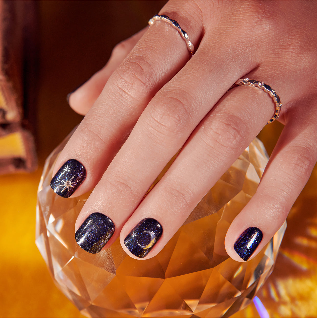 Dark sparkly nail polish with celestial designs on manicured nails, worn with silver rings.