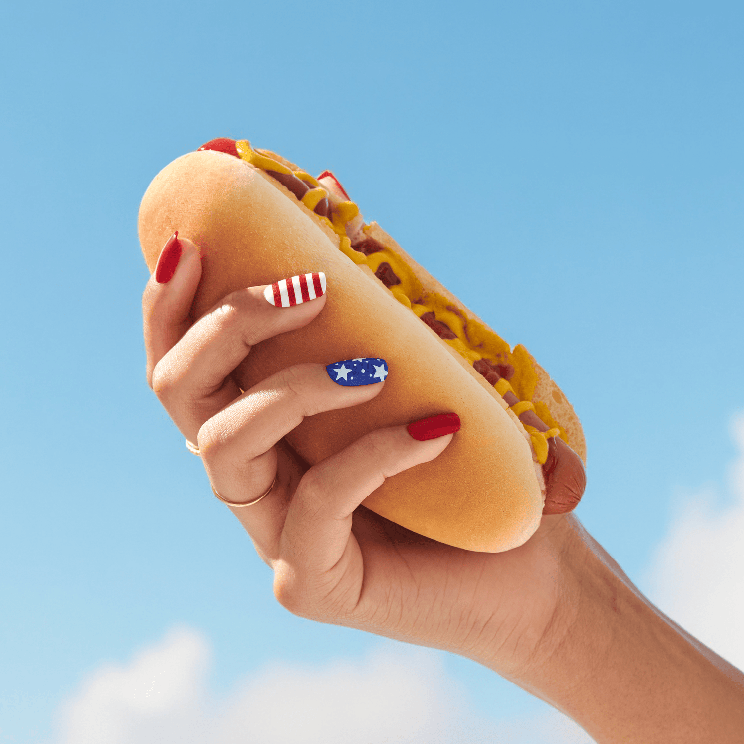 Hand with red and blue star and stripes manicure holding a hot dog with mustard against a clear blue sky