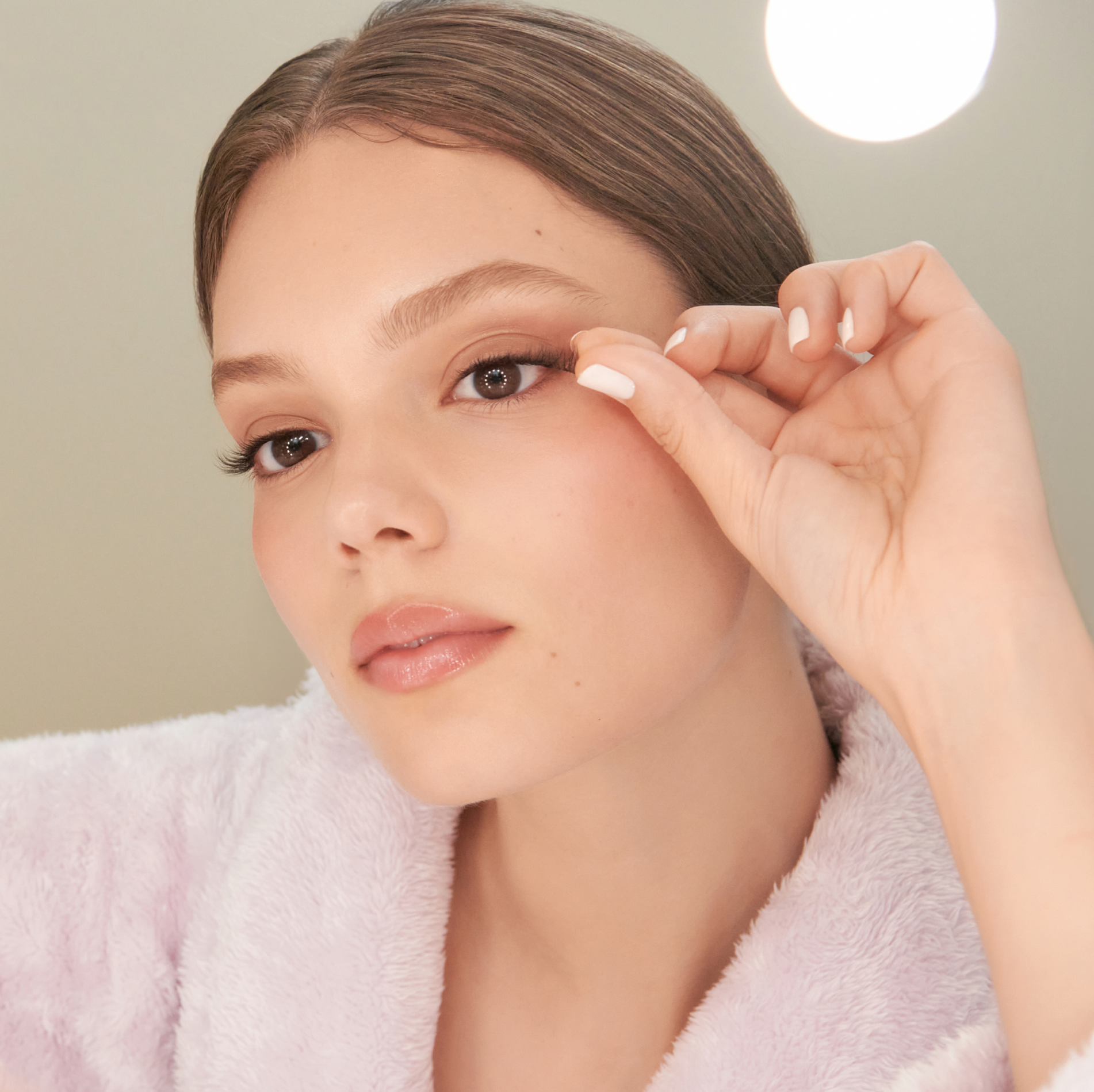 A woman looking in the mirror while applying a full strip of imPRESS press on lashes with one hand, showing just how easy it is to apply.