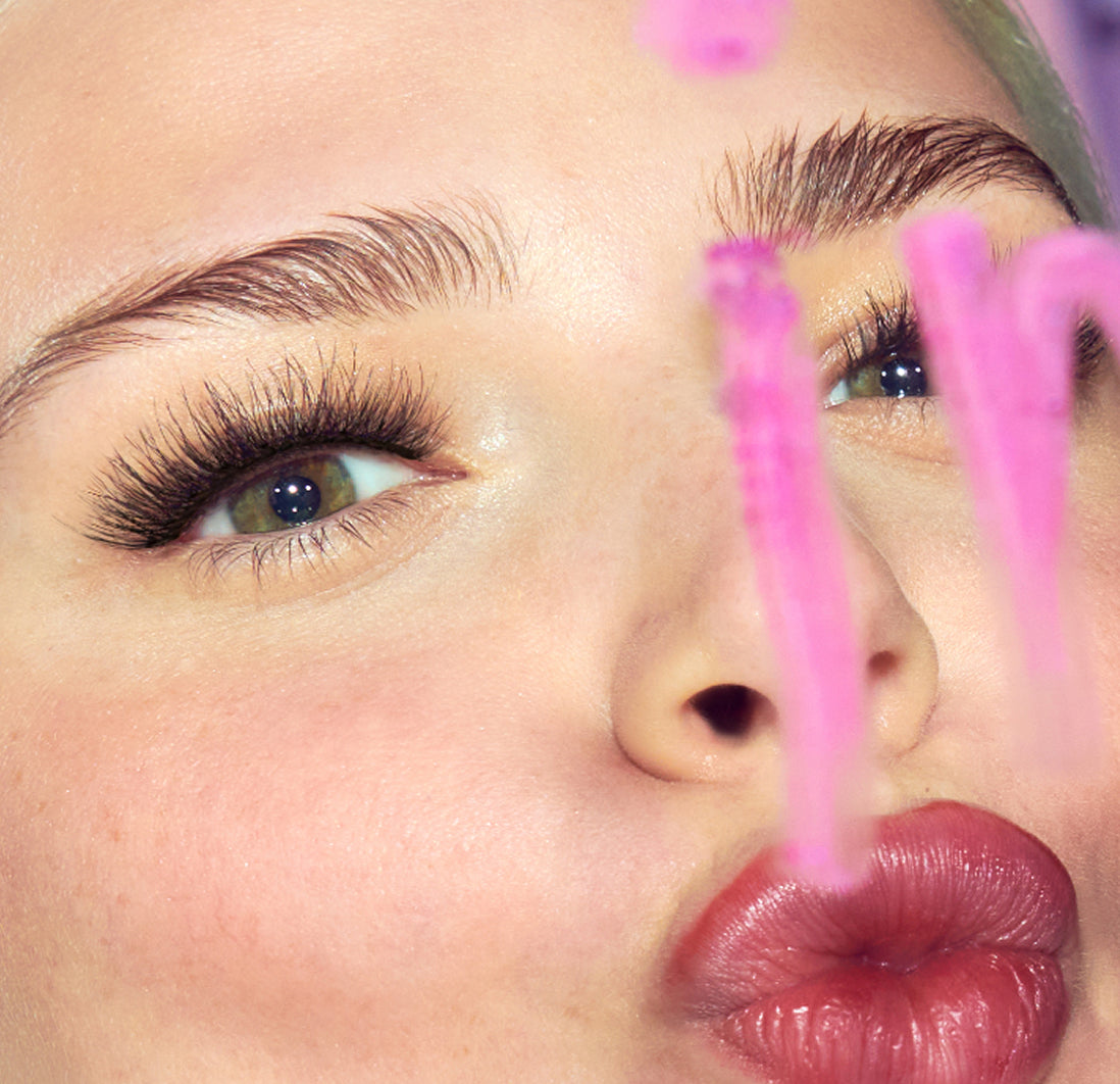Close-up of woman's face showcasing dramatic lashes, defined brows, and bold berry lips with pink filter.