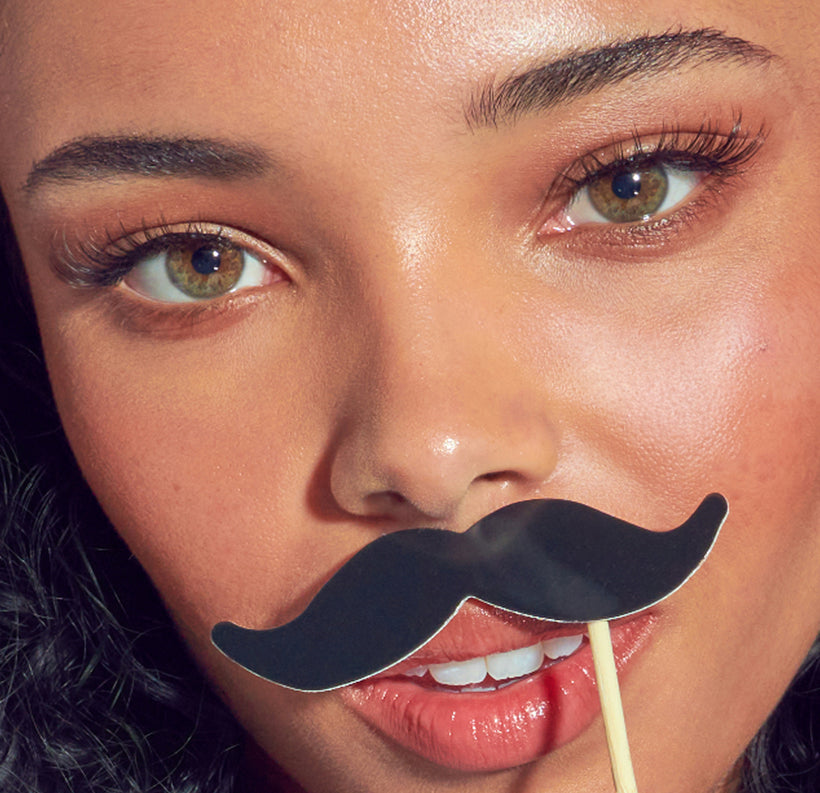 Woman holding black novelty mustache prop on stick, smiling at camera.