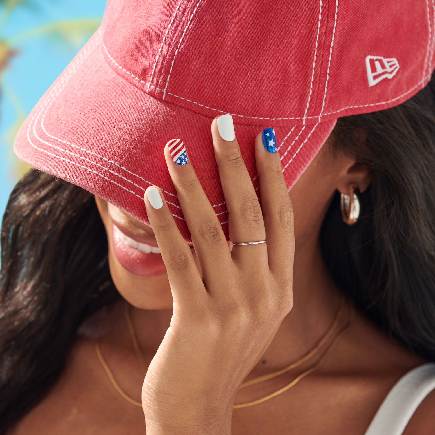 Woman wearing a red cap with a New Era logo, covering her face with her hand, showing off nail art.