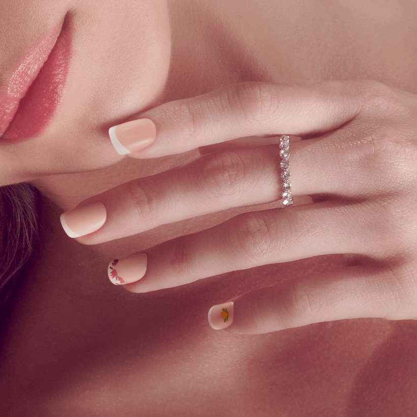 Close-up of a hand resting near a woman's face, showcasing natural pink press-on nails with white tips and small floral designs on two nails, and a silver ring with diamonds.