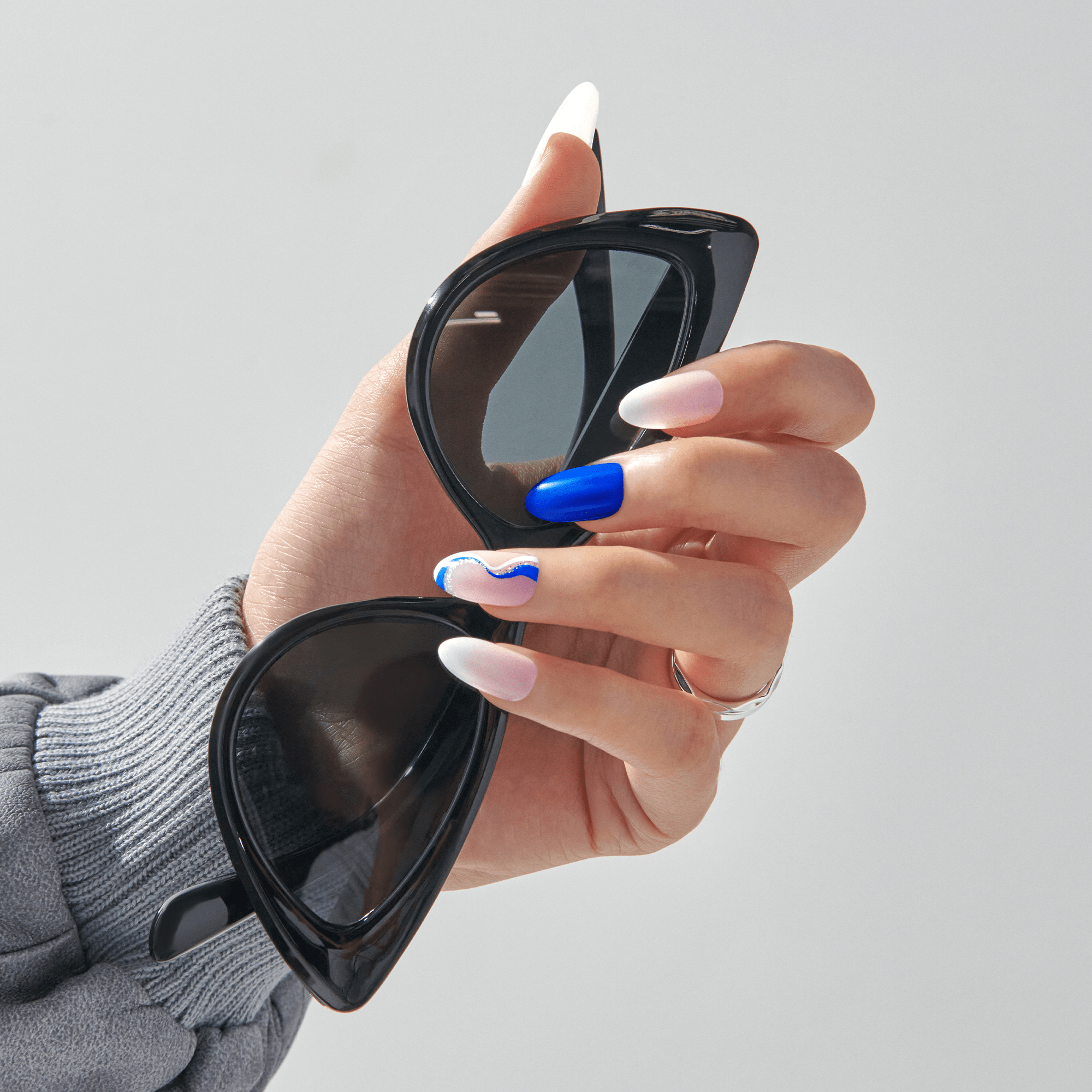 Hand holding black cat-eye sunglasses, showing press-on nails with neutral matte, bold blue, and wavy blue-and-white designs.